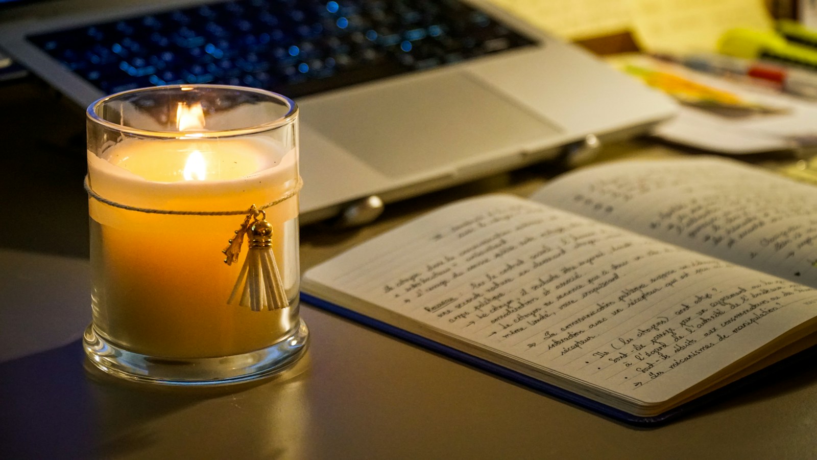 An open notebook with handwritten notes next to a candle and a laptop